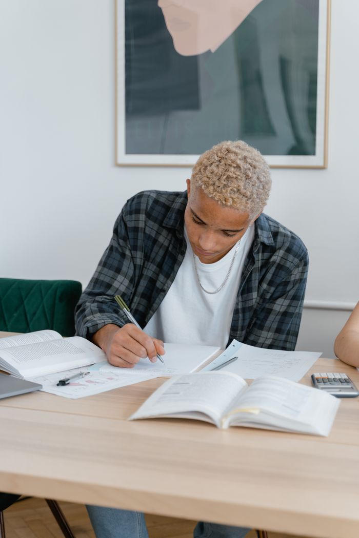 Teen student writing at home desk, surrounded by open books, engaged in study.